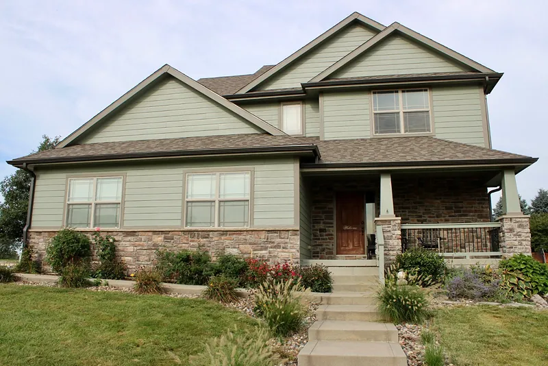 Front of a two-story Waukee home with sage green siding, tan-brown trim, stacked stone and a covered front porch after an exterior repaint by Mission Painting